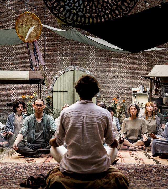 Eine Gruppe von Menschen sitzt im meditativen Schneidersitz vor einer Person, von der man nur den Rücken sieht. Alle haben die Augen geschlossen bis auf Doro Decker (Alina Levshin), die sich am Rand mit Meike (Janina Stopper) aus der Gruppe unterhält.