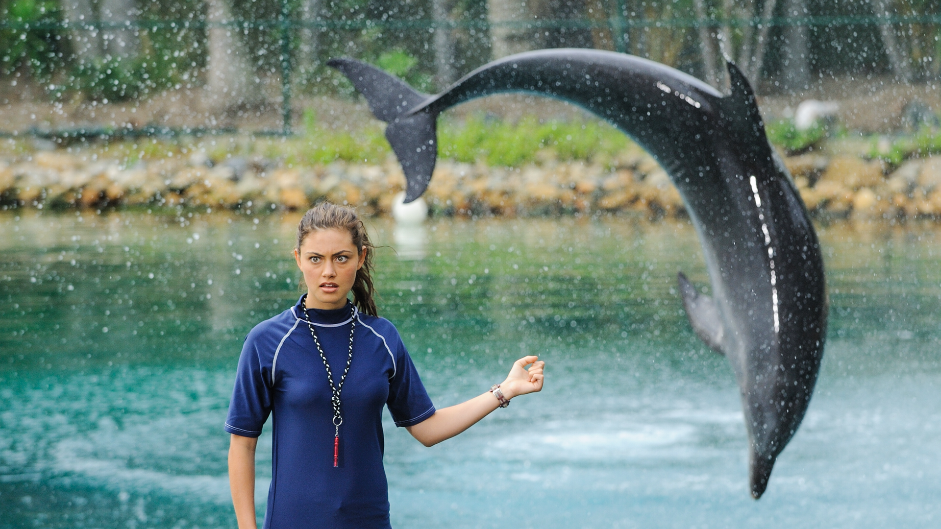 Cleo steht als Assistentin des Delfintrainers im Vordergrund und blickt verwundert nach vorne. Ihren linken Arm hält sie angewinkelt zur Seite. Im Hintergrund springt ein Delfin in einem Bogen ins Wasser.