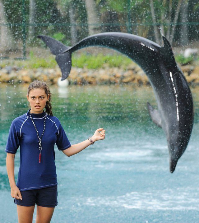 Cleo steht als Assistentin des Delfintrainers im Vordergrund und blickt verwundert nach vorne. Ihren linken Arm hält sie angewinkelt zur Seite. Im Hintergrund springt ein Delfin in einem Bogen ins Wasser.