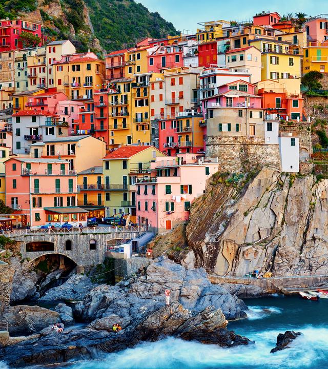 Blick vom Meer aus auf ein Dorf, auf Manarola. Bunte Häuser schmiegen sich an eine steile, felsige Küste.