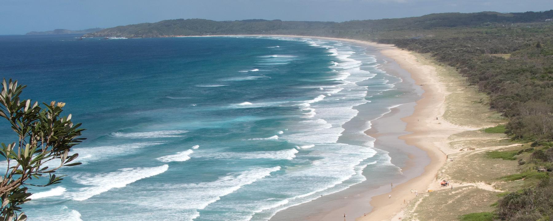 Panoramaaufnahme eines weitläufigen Sandstrandes der in ein dichtes Waldgebiet übergeht. Am blauem Himmel hängen einige Wolken.  