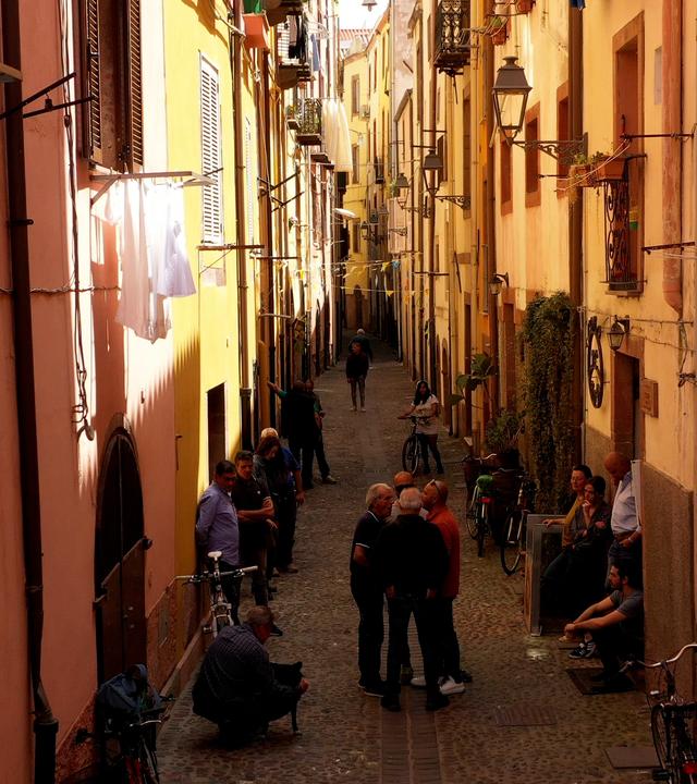 Enge Altstadtgasse im Dorf Bosa mit ockergelben Häuserwänden und einigen Menschen.