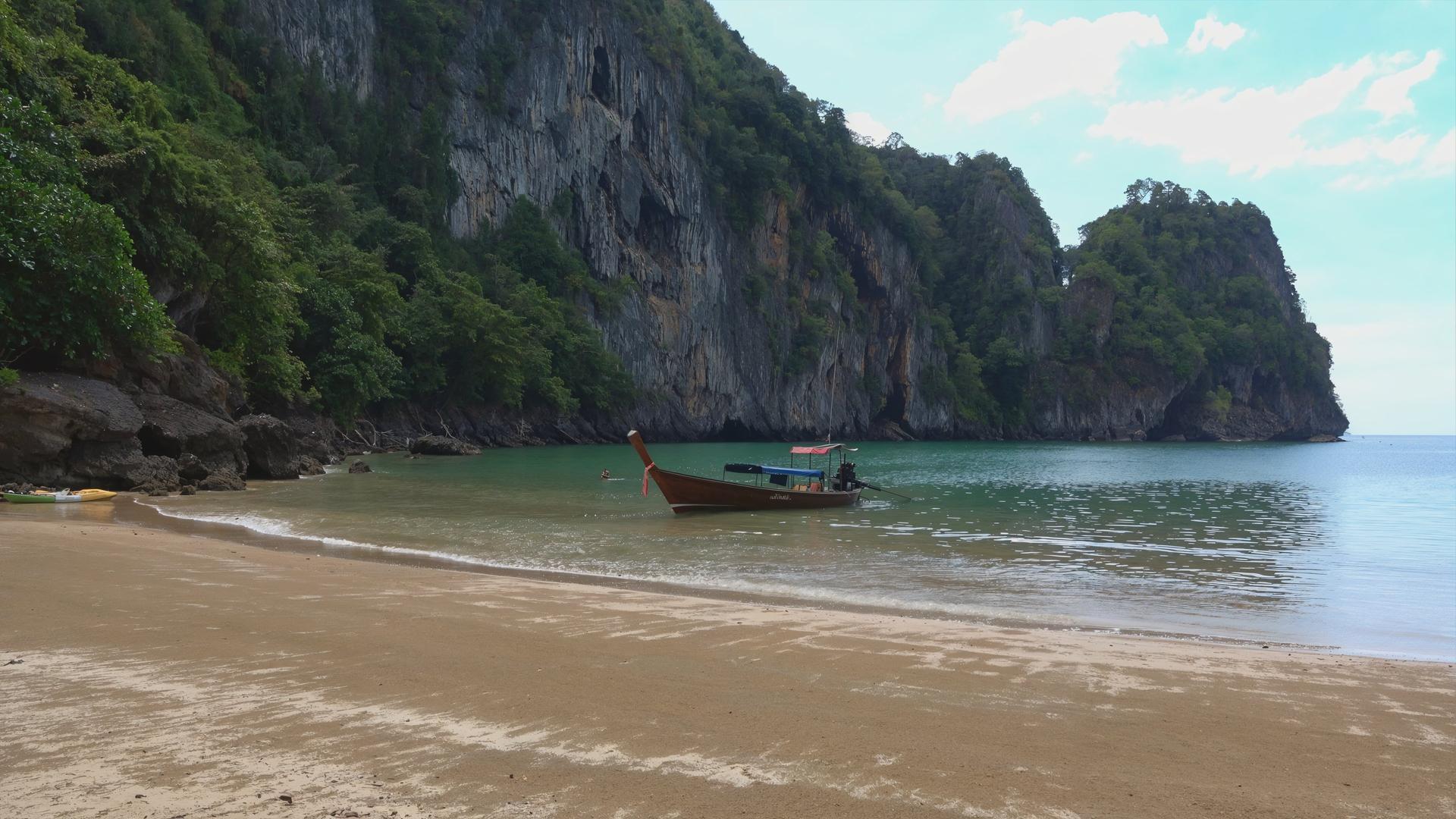 Sandstrand, an dem ein Longtailboot liegt, dahinter fällt eine spärlich bewachsene Felswand steil ins Meer hinab.