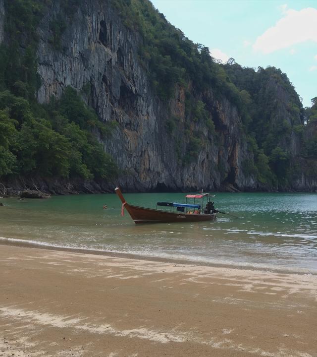Sandstrand, an dem ein Longtailboot liegt, dahinter fällt eine spärlich bewachsene Felswand steil ins Meer hinab.
