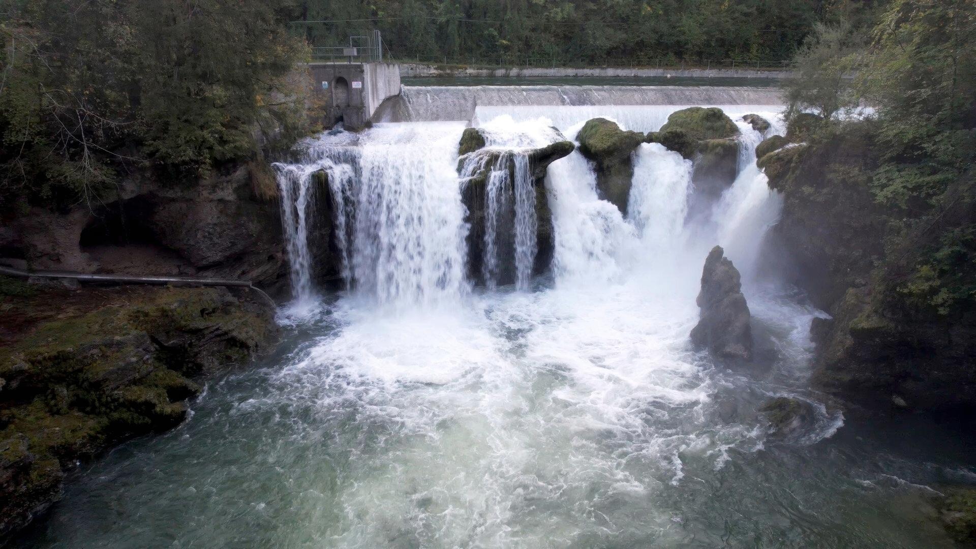 Das Bild zeigt einen beeindruckenden Wasserfall, der Teil des Flusses Traun in Österreich ist. Der Wasserfall stürzt aus mehreren Stufen herunter und bildet dabei schäumendes Wasser, das in den darunterliegenden Fluss fließt. Am oberen Rand des Wasserfalls ist eine Betonmauer sichtbar, die möglicherweise eine Wehranlage darstellt. Um den Wasserfall herum sind natürliche Felsen und flussnahe Pflanzen zu sehen. Im Hintergrund sind Wälder sichtbar, die sich an den Ufern des Flusses erstrecken. Die gesamte Szenerie wirkt dynamisch und lebendig, geprägt von dem rauschenden Wasser und der umgebenden Natur.