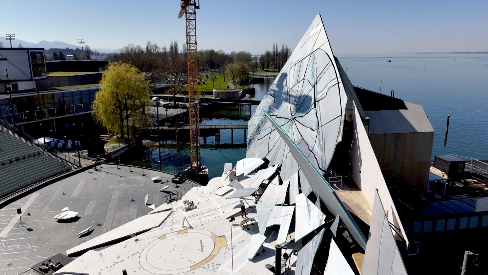 Das Bild zeigt den Aufbau des Bühnenbildes für die Bregenzer Festspiele, speziell für die Aufführung von Verdis "La traviata". Im Vordergrund ist eine imposante, moderne Bühnenkonstruktion zu sehen, die aus großen, reflektierenden Flächen in Form eines spitzen Dreiecks besteht. Diese Elemente sind mit einem gebrochenen Design versehen, das an zerbrochenes Glas erinnert.   Im Hintergrund erstreckt sich das Wasser des Bodensees, und entlang des Ufers sind Bäume und eine Rasenfläche sichtbar. Ein Kran ist zu sehen, der Materialien für den Bühnenaufbau anhebt. In der unteren linken Ecke des Bildes sind einige Bauarbeiten und kleinere Elemente auf dem Boden platziert.   Das Bild vermittelt einen Eindruck von der Dynamik und der technischen Komplexität, die mit der Aufführung auf der größten Seebühne der Welt verbunden sind. Der Himmel ist klar und sonnig, was die Szenerie hell und einladend erscheinen lässt.