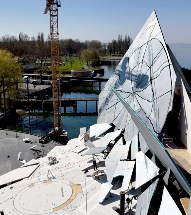 Das Bild zeigt den Aufbau des Bühnenbildes für die Bregenzer Festspiele, speziell für die Aufführung von Verdis "La traviata". Im Vordergrund ist eine imposante, moderne Bühnenkonstruktion zu sehen, die aus großen, reflektierenden Flächen in Form eines spitzen Dreiecks besteht. Diese Elemente sind mit einem gebrochenen Design versehen, das an zerbrochenes Glas erinnert.   Im Hintergrund erstreckt sich das Wasser des Bodensees, und entlang des Ufers sind Bäume und eine Rasenfläche sichtbar. Ein Kran ist zu sehen, der Materialien für den Bühnenaufbau anhebt. In der unteren linken Ecke des Bildes sind einige Bauarbeiten und kleinere Elemente auf dem Boden platziert.   Das Bild vermittelt einen Eindruck von der Dynamik und der technischen Komplexität, die mit der Aufführung auf der größten Seebühne der Welt verbunden sind. Der Himmel ist klar und sonnig, was die Szenerie hell und einladend erscheinen lässt.