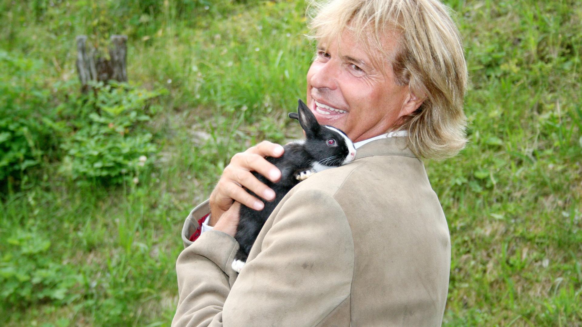 Das Bild zeigt einen Mann mit langen, blonden Haaren, der lächelt und einen kleinen schwarzen Hasen mit weißen Abzeichen auf seinem Arm trägt. Er trägt einen hellen Anzug mit einer roten Krawatte. Der Hintergrund besteht aus grünen Wiesen und Büschen, was auf eine ländliche Umgebung hinweist. Die Stimmung wirkt freundlich und entspannt.