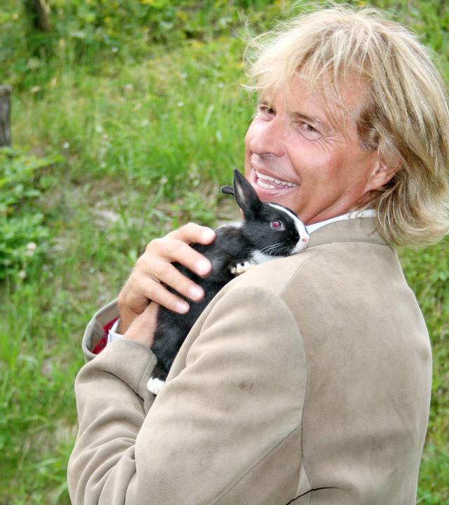 Das Bild zeigt einen Mann mit langen, blonden Haaren, der lächelt und einen kleinen schwarzen Hasen mit weißen Abzeichen auf seinem Arm trägt. Er trägt einen hellen Anzug mit einer roten Krawatte. Der Hintergrund besteht aus grünen Wiesen und Büschen, was auf eine ländliche Umgebung hinweist. Die Stimmung wirkt freundlich und entspannt.