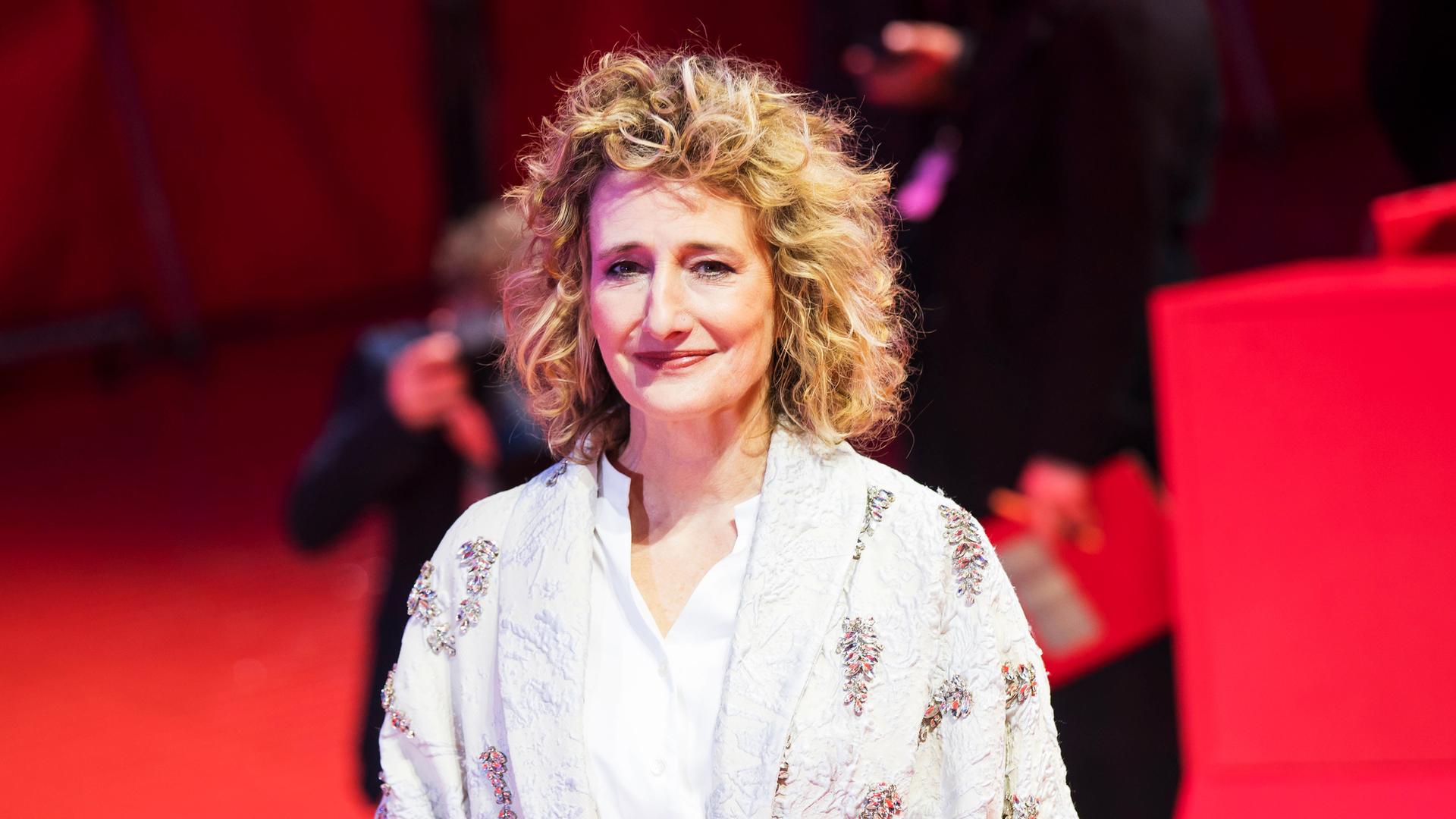 Eine Frau (Tricia Tuttle) mit blonden lockigen Haaren, die eine Bluse und einen Mantel trägt. Sie steht auf einem roten Teppich und hinter ihr sind Fotografen.