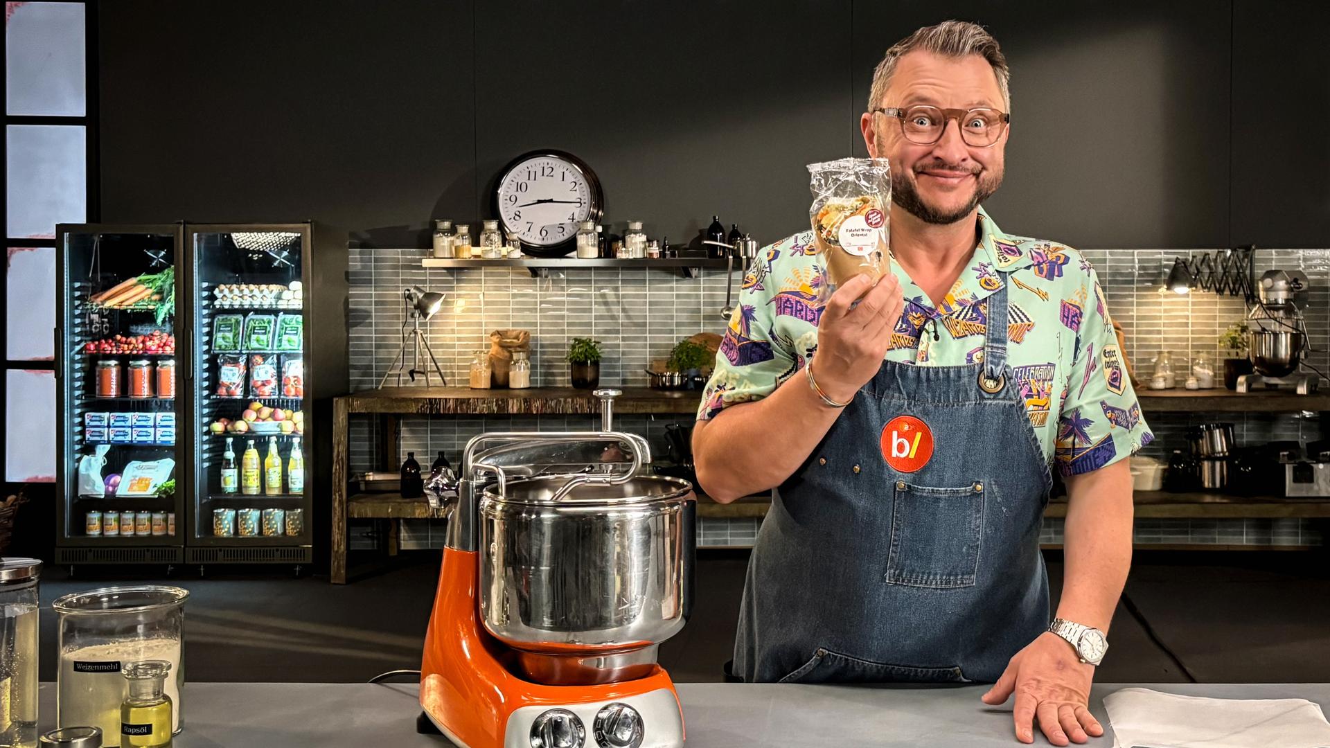 Sebastian Lege steht an der Arbeitsfläche seiner Werkstattküche. Er hält ein verpacktes Falafel-Sandwich in der Hand.