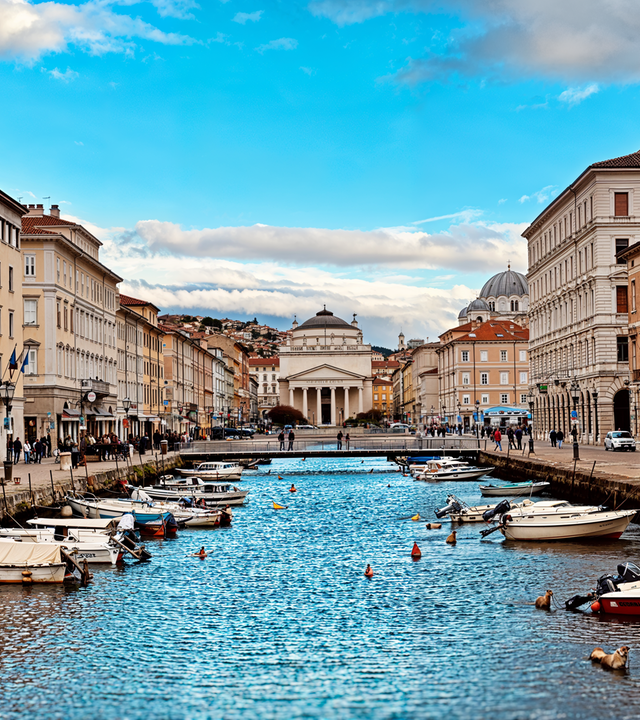 Blick entlang des Canale Grande in Triest mit Booten, historischen Gebäuden und einer Kirche im Hintergrund.