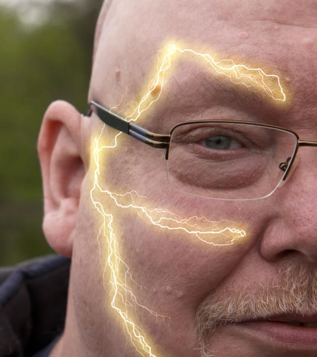 Ein Mann mittleren Alters mit Brille schaut in die Kamera. Auf der linken Seite seines Gesichts verlaufen gelb leuchtenden Linien. Sie symbolisieren den Trigeminusnerv mit seinen drei Ästen.