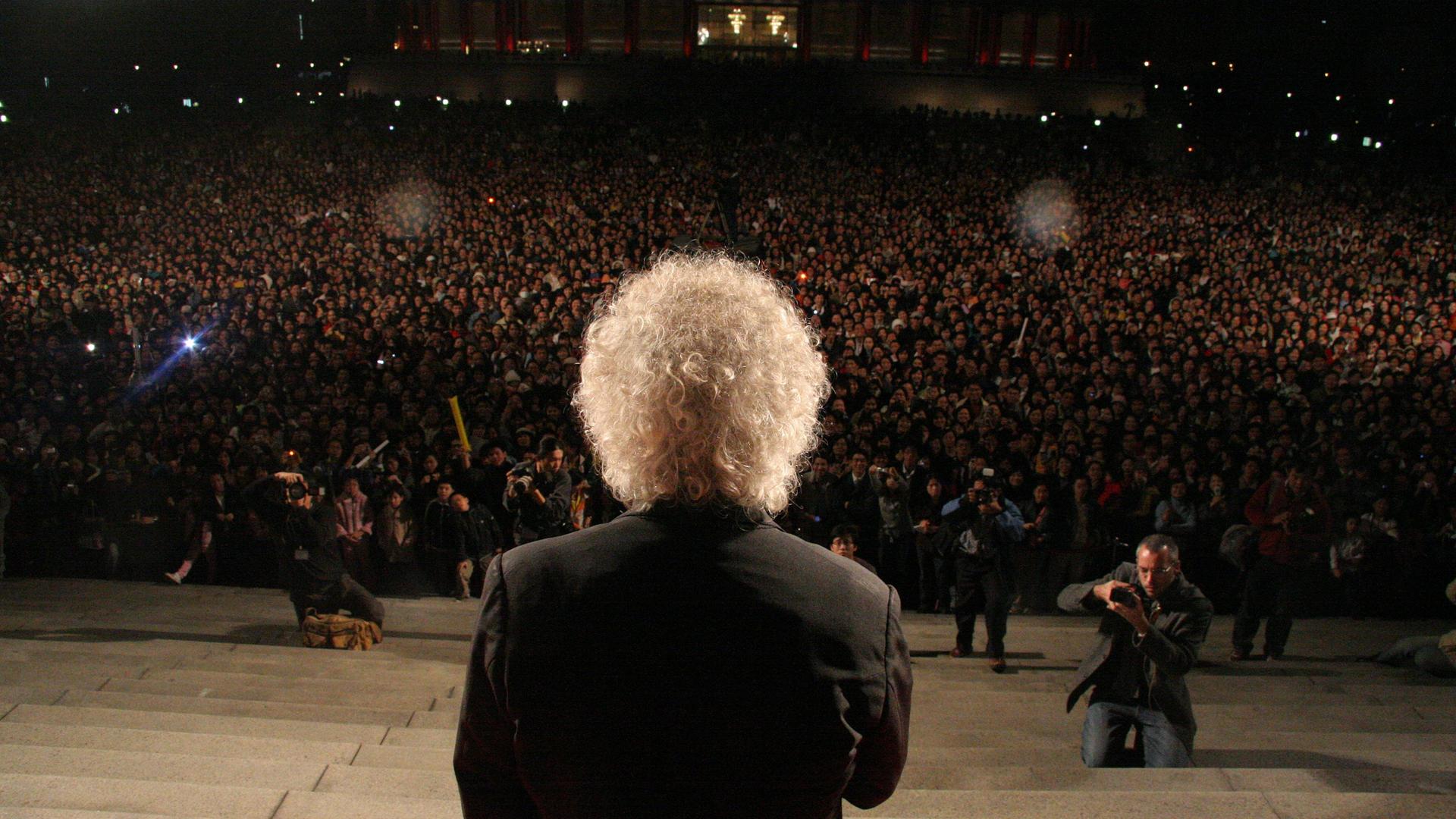 Dirigent Sir Simon Rattle steht mit dem Rücken zur Kamera auf einer Treppe. Am Fuß der Treppe ist eine Menschenmasse sowie Fotografen zu sehen.