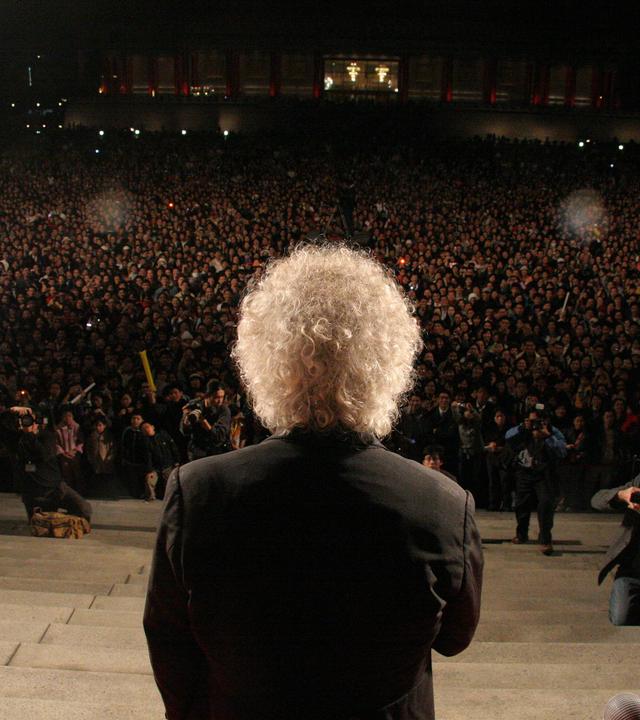 Dirigent Sir Simon Rattle steht mit dem Rücken zur Kamera auf einer Treppe. Am Fuß der Treppe ist eine Menschenmasse sowie Fotografen zu sehen.
