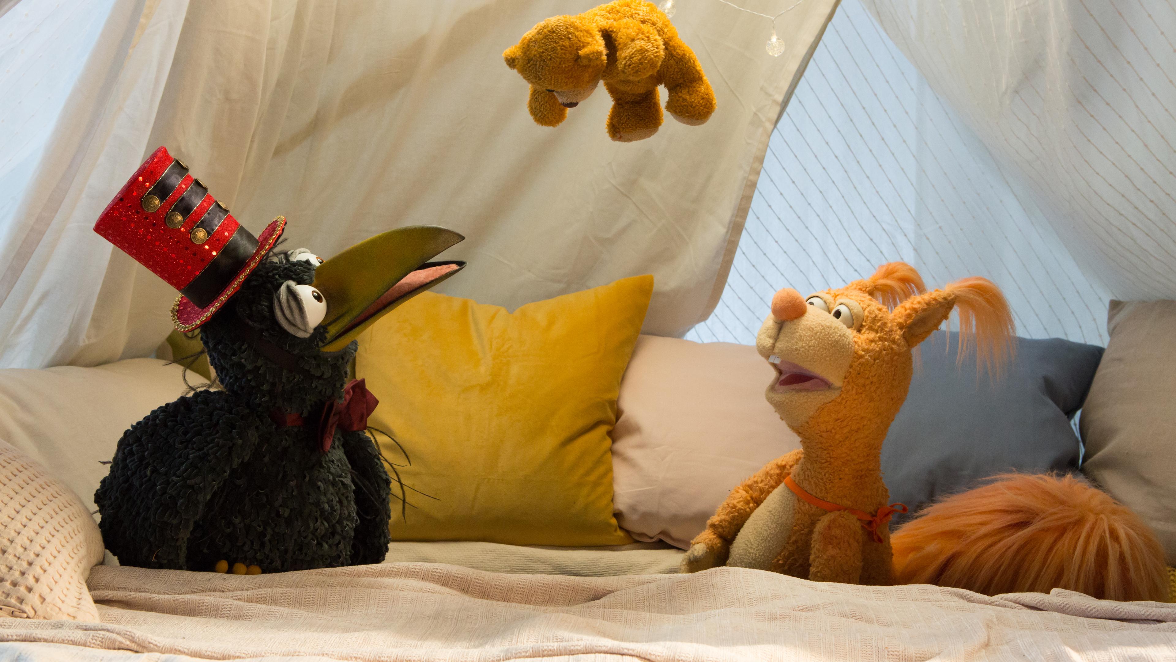 Rudi und Ella hocken in der Kuschelecke und schauen staunend nach oben: Über ihnen hängt Teddy an einem Faden. Rudi trägt einen Zylinder wie ein Zirkusdirektor.