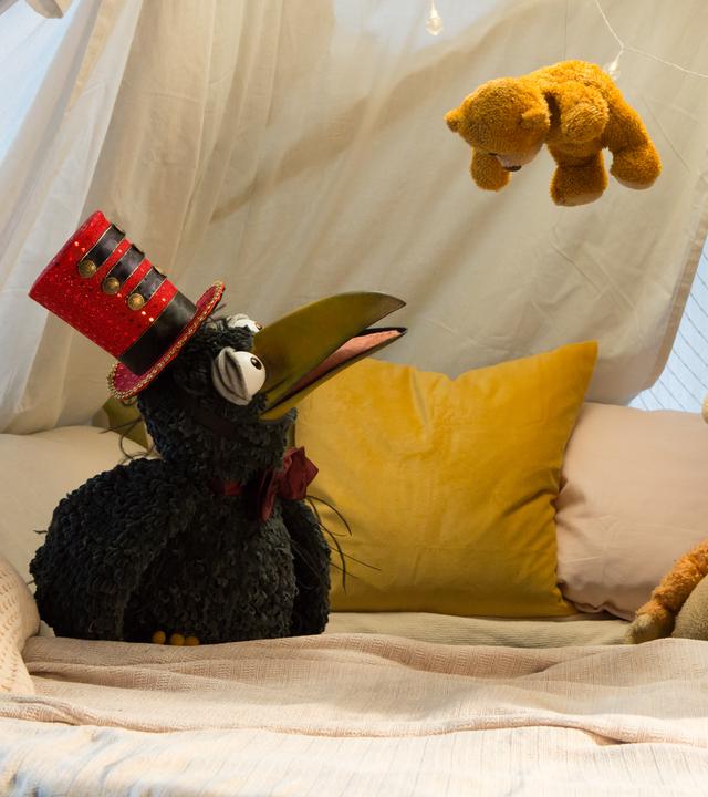 Rudi und Ella hocken in der Kuschelecke und schauen staunend nach oben: Über ihnen hängt Teddy an einem Faden. Rudi trägt einen Zylinder wie ein Zirkusdirektor.