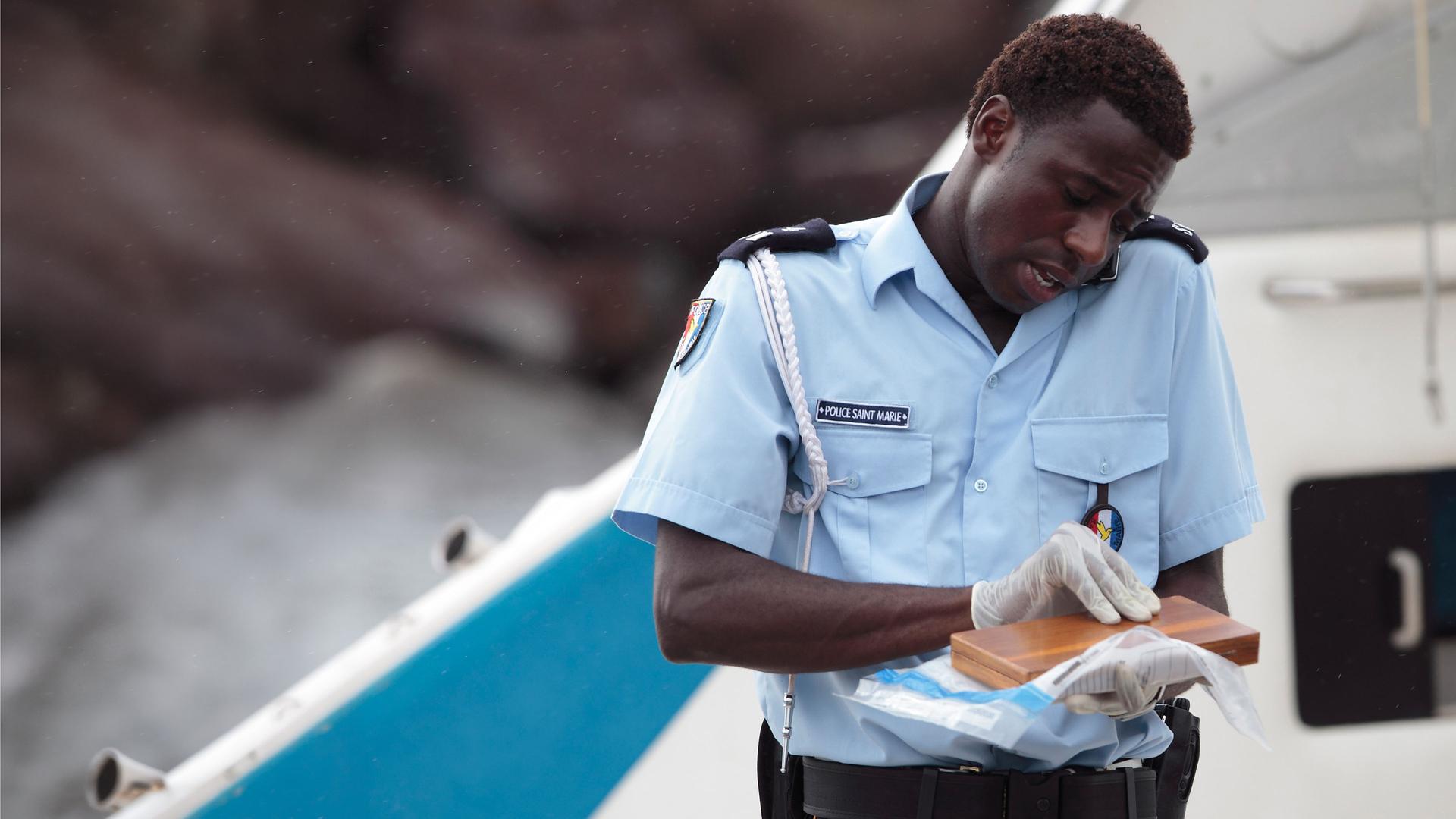 "Death in Paradise - Tropisches Fieber": Nahaufnahme Fidel (Gary Carr) auf einem Boot - er hält ein Handy zwischen Schulter und Kopf gepresst. In den Händen hält er eine kleine Holzschachtel.