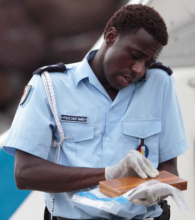 "Death in Paradise - Tropisches Fieber": Nahaufnahme Fidel (Gary Carr) auf einem Boot - er hält ein Handy zwischen Schulter und Kopf gepresst. In den Händen hält er eine kleine Holzschachtel.