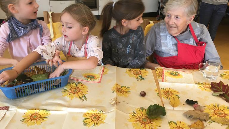  "plan b: Trubel im Altenheim - Wie Kinder und Senioren voneinander profitieren": Drei Kinder sitzen mit einer älteren Dame an einem Tisch und unterhalten sich.