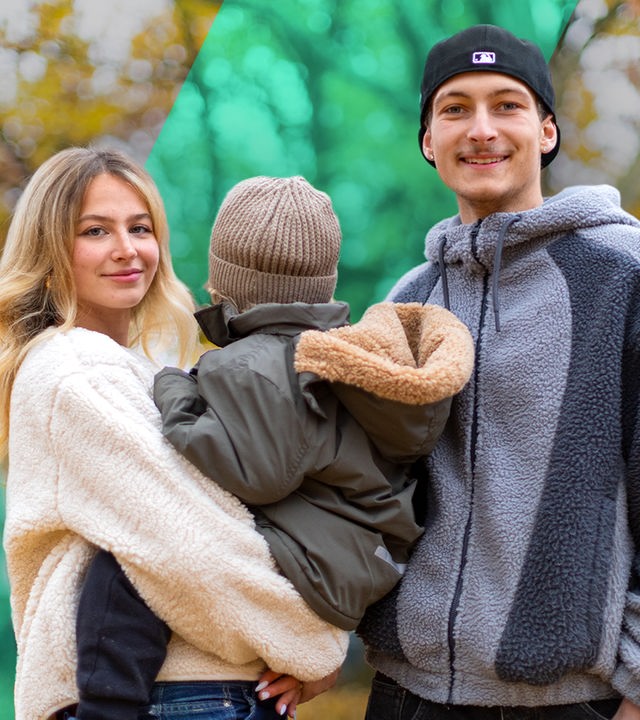 Eine junge Frau und ein junger Mann lächeln. Er trägt eine Mütze. Sie trägt ein kleines Kind auf dem Arm, das mit dem Gesicht nach hinten schaut.