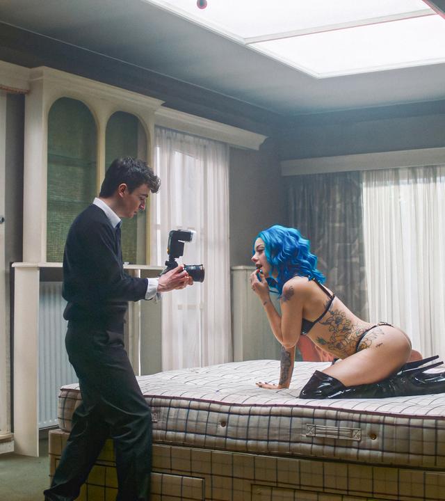 Eine Frau mit blauen Haaren und in schwarzer Unterwäsche räkelt sich aur einem Bett, während ein junger Mann sie mit einer Kamera filmt.
