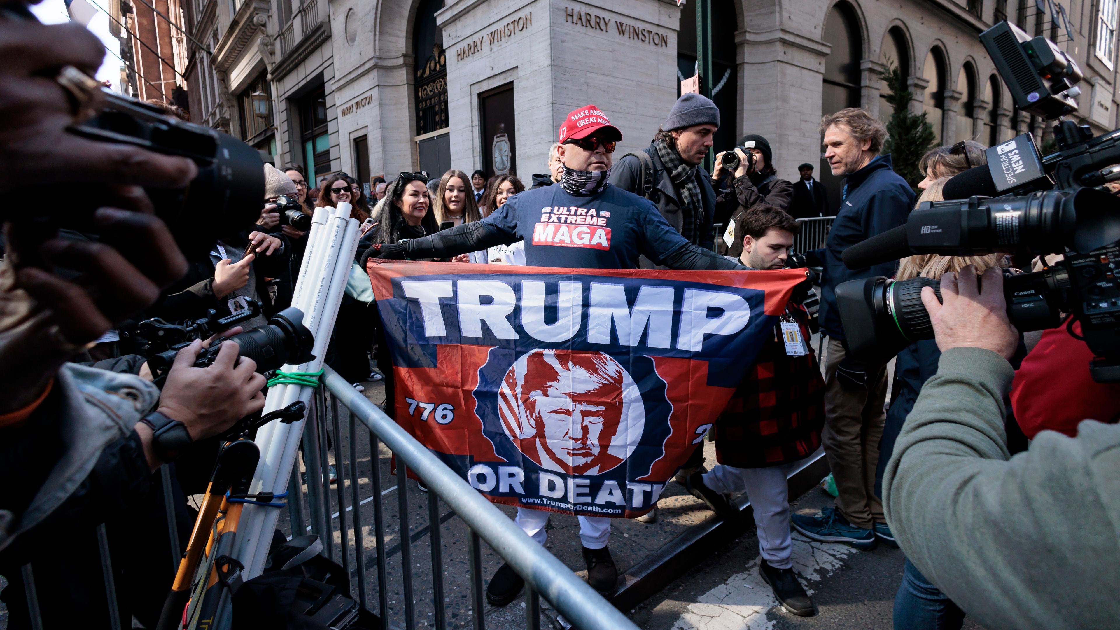 Ein Frau steht zwischen mehreren anderen Personen auf der Straße und hält eine Flagge mit der Aufschrift "Trump" in den Händen.