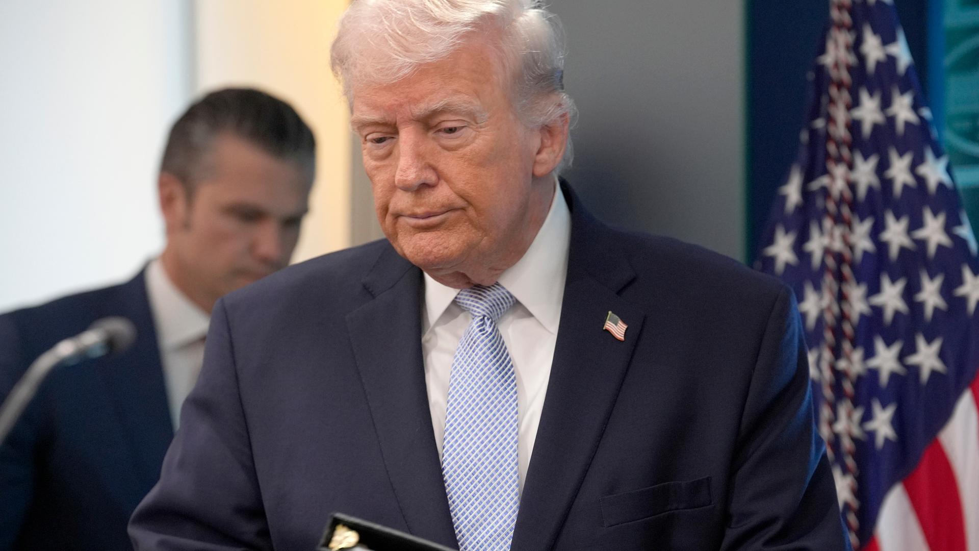 US-Präsident Donald Trump im Presseraum des Weißen Hauses mit gesenktem Blick. Im Hintergrund steht Kriegsminister Hegseth.