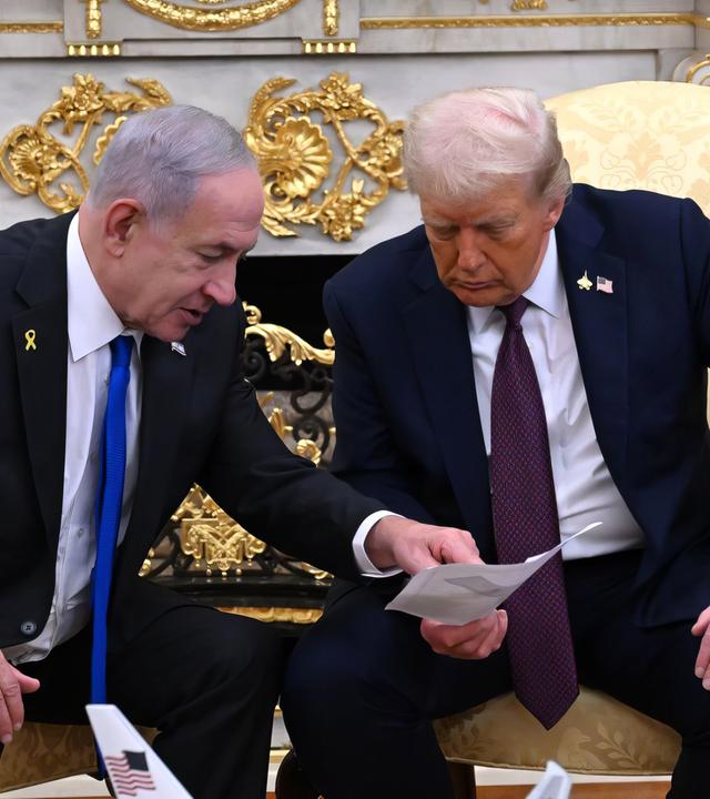 Zwei ältere Männer in dunklen Anzügen sitzen auf goldenen Stühlen und beugen sich über ein Blatt Papier, das einer in der Hand hält.