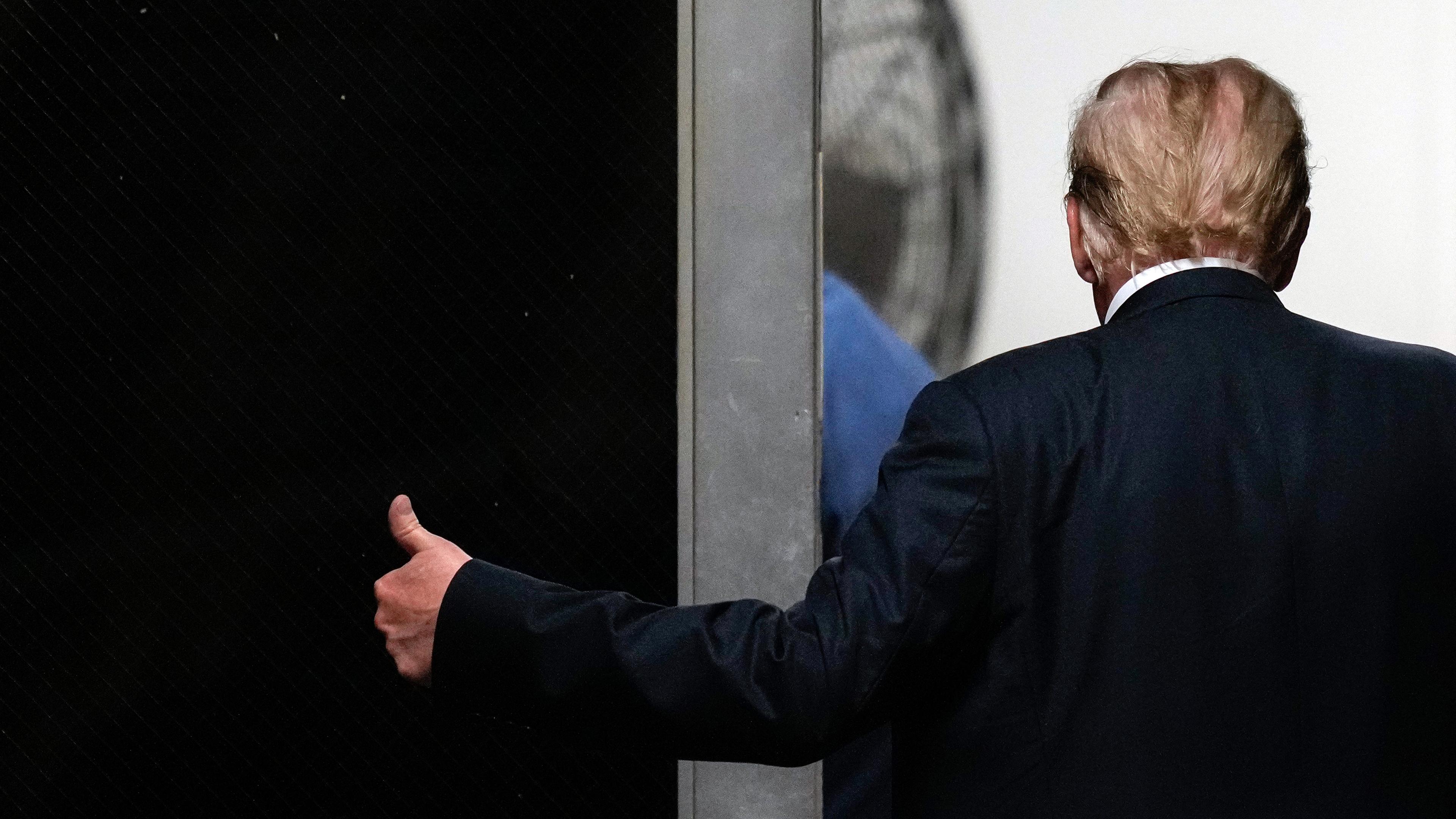 Donald Trump ist von hinten zu sehen, wie er mit der linken Hand einen Daumen nach oben zeigt. 