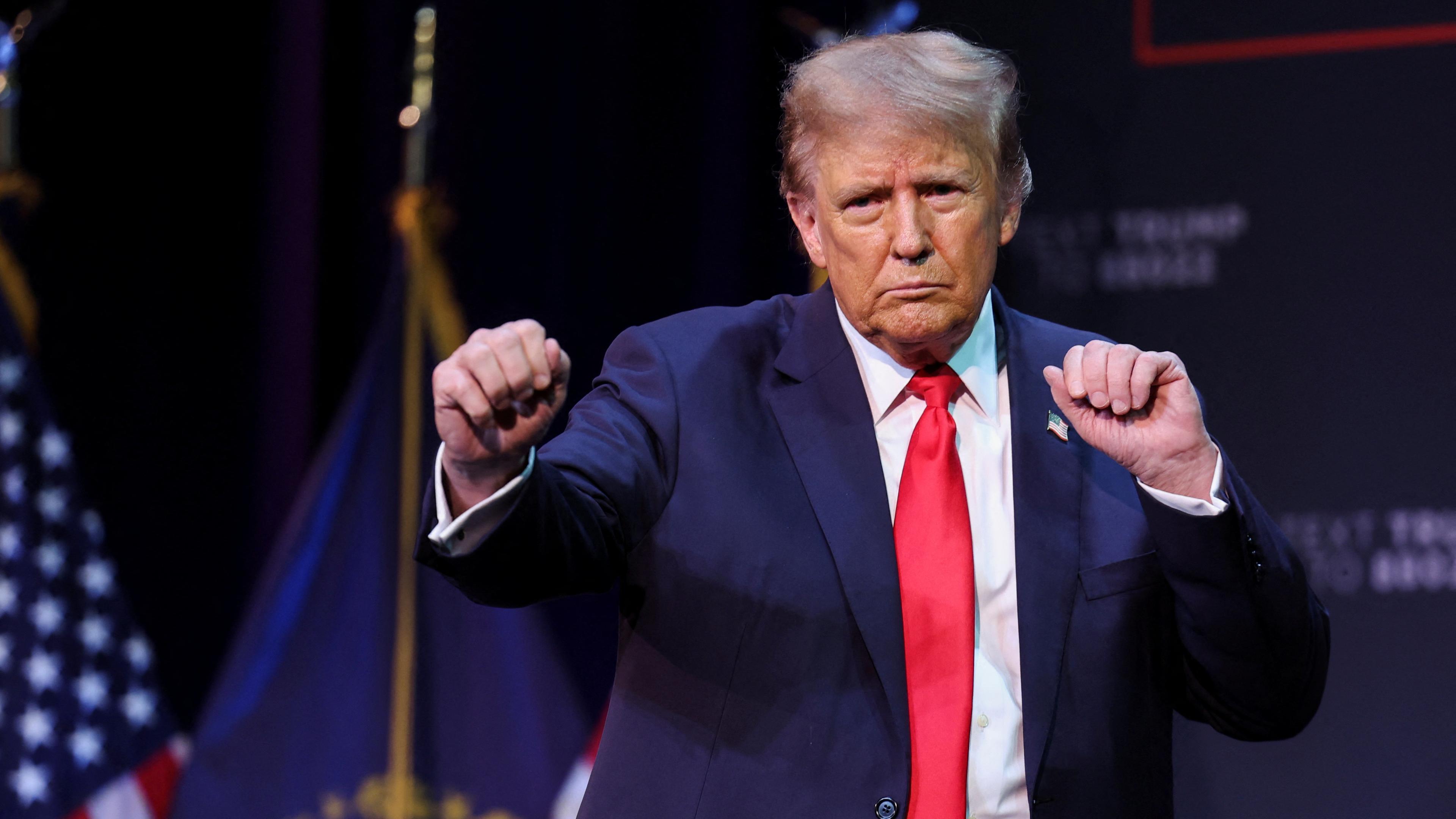 Der ehemalige US-President Trump tanzt mit den Händen zu Fäusten geballt auf einer Bühne, im Hintergrund sind US-Flaggen.