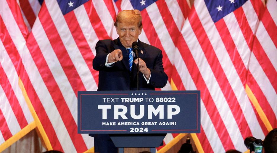 Donald Trump steht auf einer Bühne an einem Podium, hinter ihm ist die US-amerikanische Flagge zu sehen. Er zeigt in die Menge und lächelt. Auf dem Podium steht Text Trump to 8802, Trump, Make America Great Again, 2024.