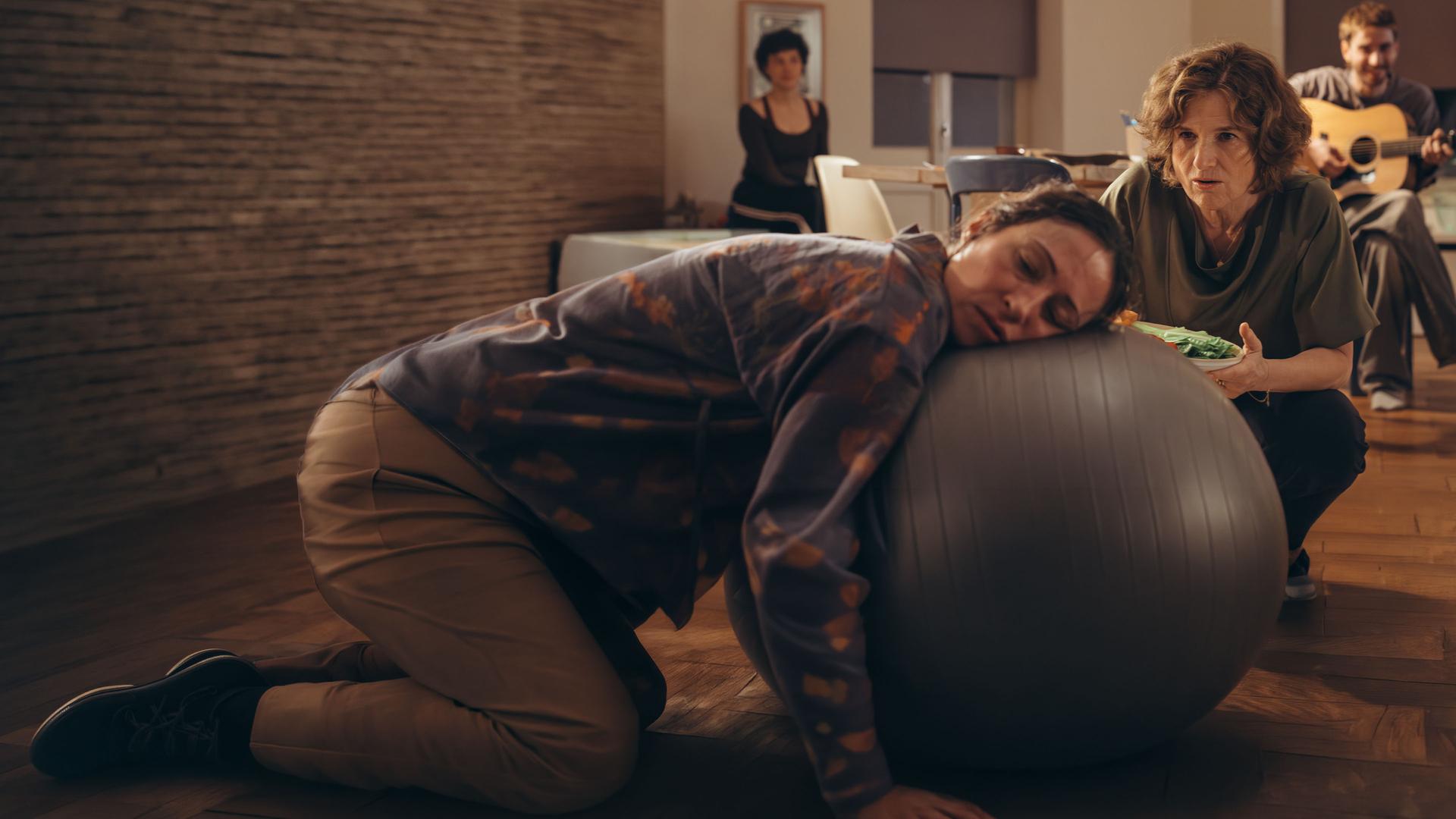 "PUSH - Trust the process": Raquel (Aenne Schwarz) liegt erschöpft mit ihrem Kopf auf einem Gymnastikball. Marcela (Victoria Trauttmansdorff) kniet neben ihr mit einem Gemüseteller und spricht mit Raquel. Leandro (James Newton) sitzt dahinter und spielt Gitarre. Greta (Lydia Amasko) sitzt im Hintergrund.