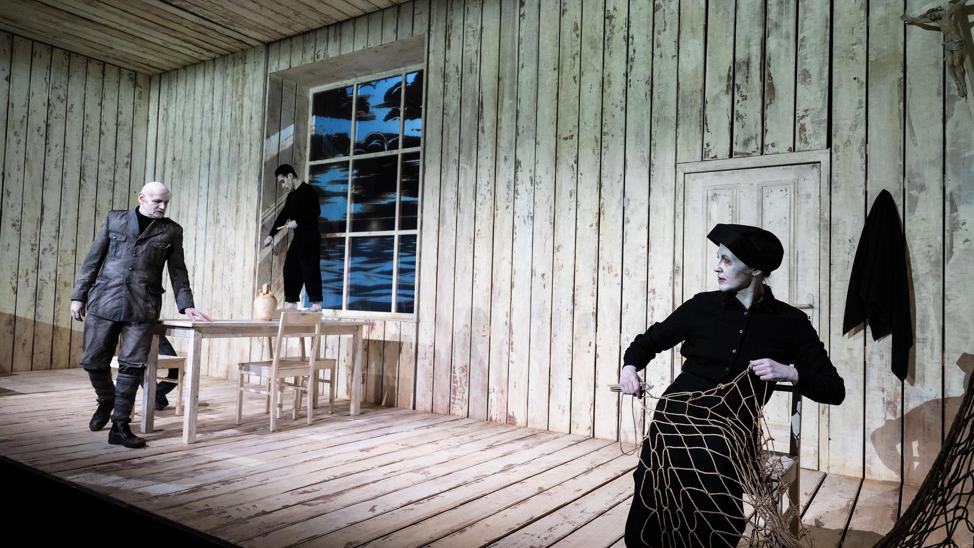Theaterszene in einer hölzernen Hütte mit drei Schauspielenden. Links geht ein älterer Mann in einer Militäruniform am Tisch vorbei. Ein jüngerer Mann in schwarzer Kleidung steht barfuß auf dem Fensterbrett und schnitzt. Rechts im Vordergrund sitzt eine Frau in schwarzer Kleidung mit Kopfbedeckung auf einem Stuhl und arbeitet an einem großen Fischernetz. 