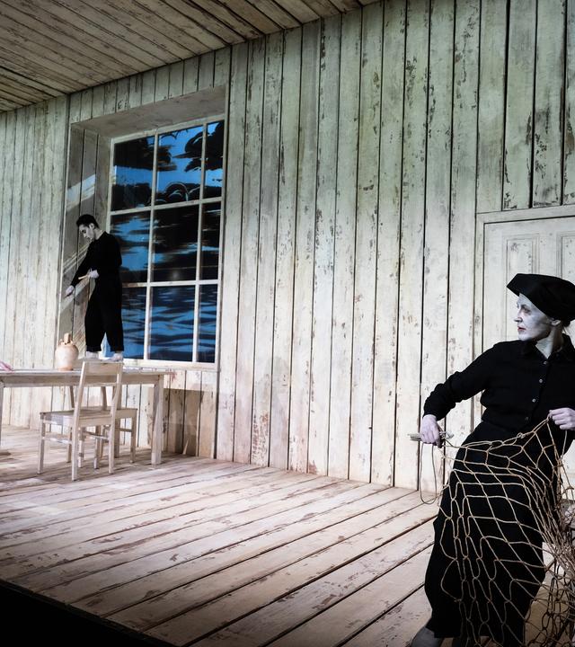 Theaterszene in einer hölzernen Hütte mit drei Schauspielenden. Links geht ein älterer Mann in einer Militäruniform am Tisch vorbei. Ein jüngerer Mann in schwarzer Kleidung steht barfuß auf dem Fensterbrett und schnitzt. Rechts im Vordergrund sitzt eine Frau in schwarzer Kleidung mit Kopfbedeckung auf einem Stuhl und arbeitet an einem großen Fischernetz. 