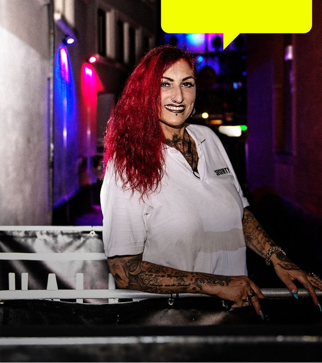 Anke Henkel, eine Frau mit langen roten Haaren und vielen Tattoos, blickt freundlich in die Kamera. Sie steht am Eingang der Discothek "Markthalle", einem Klub in Kaiserslautern.