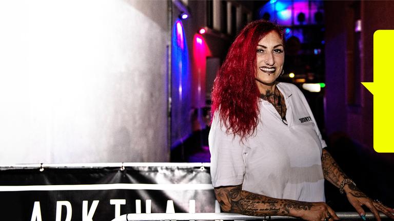 Anke Henkel, eine Frau mit langen roten Haaren und vielen Tattoos, blickt freundlich in die Kamera. Sie steht am Eingang der Discothek "Markthalle", einem Klub in Kaiserslautern.