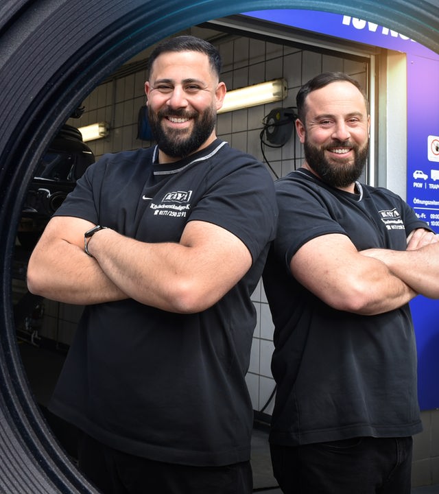 Serkan und Engin Kaya stehen lachend, mit verschränkten Armen und Schulter an Schulter vor einem geöffneten Werkstatttor. Im Hintergrund steht ein Auto auf einer Hebebühne.
