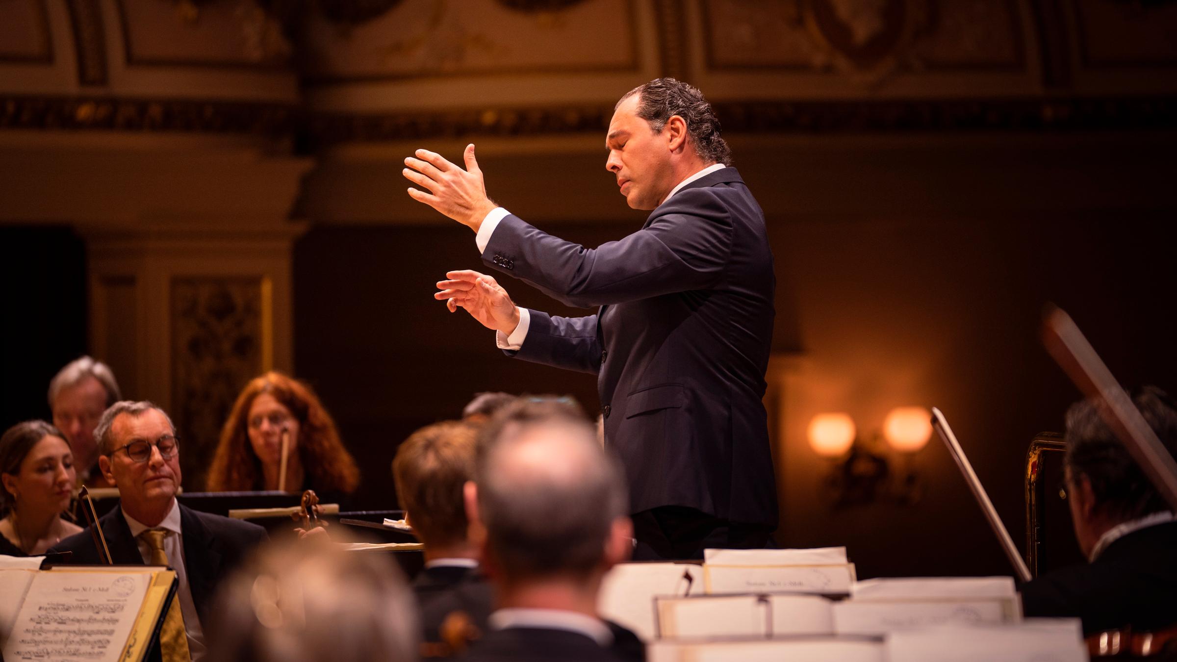 Seitlicher Blick auf den Dirigenten Tugan Sokhiev. Er dirigiert und hat dabei die Augen geschlossen. Um ihn herum sitzen die Orchestermusiker.