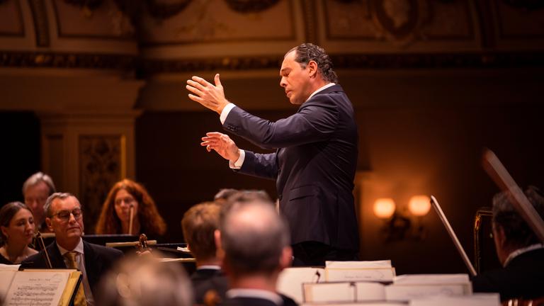 Seitlicher Blick auf den Dirigenten Tugan Sokhiev. Er dirigiert und hat dabei die Augen geschlossen. Um ihn herum sitzen die Orchestermusiker.
