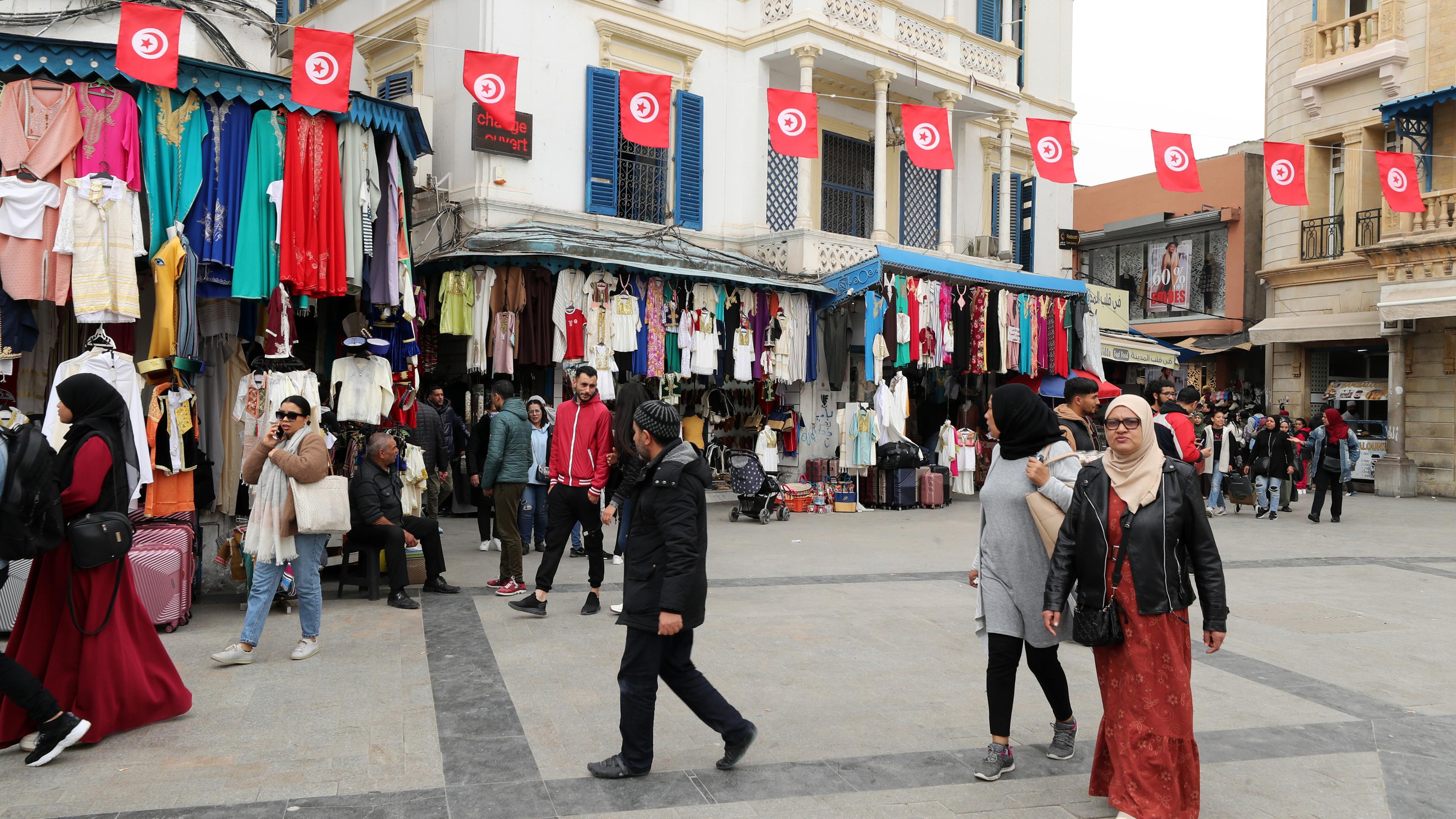 Eine belebte Straße mit Marktsänden in Tunesien. Über der Straße hängt eine Girlande aus der tunesischen Flagge.