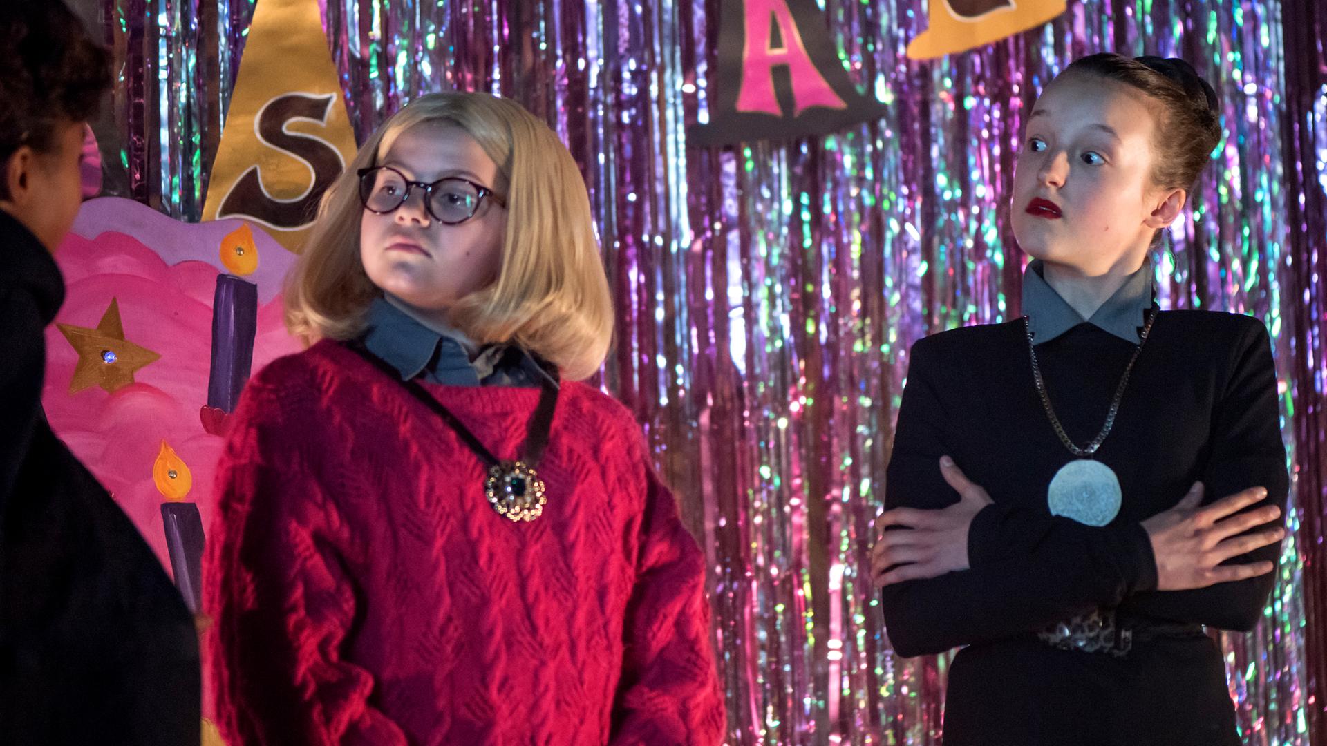 Mildred und Maude stehen als Frau Graustein und Frau Harschmann verkleidet auf der Bühne. Mildred hat einen sehr strengen Blick.