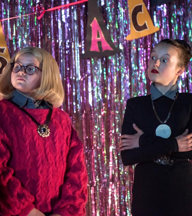 Mildred und Maude stehen als Frau Graustein und Frau Harschmann verkleidet auf der Bühne. Mildred hat einen sehr strengen Blick.