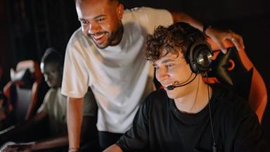Drei junge Männer spielen gemeinsam in einem abgedunkelten Raum am Computer. Im Vordergrund trägt einer von ihnen ein Headset mit Mikrofon, während ein Freund ihm lächelnd über die Schulter schaut. Die Szene steht sinnbildlich für die ursprünglich stark vom Gaming geprägte Twitch-Community.