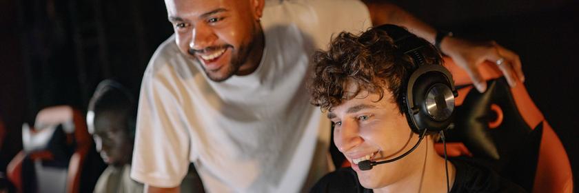 Drei junge Männer spielen gemeinsam in einem abgedunkelten Raum am Computer. Im Vordergrund trägt einer von ihnen ein Headset mit Mikrofon, während ein Freund ihm lächelnd über die Schulter schaut. Die Szene steht sinnbildlich für die ursprünglich stark vom Gaming geprägte Twitch-Community.
