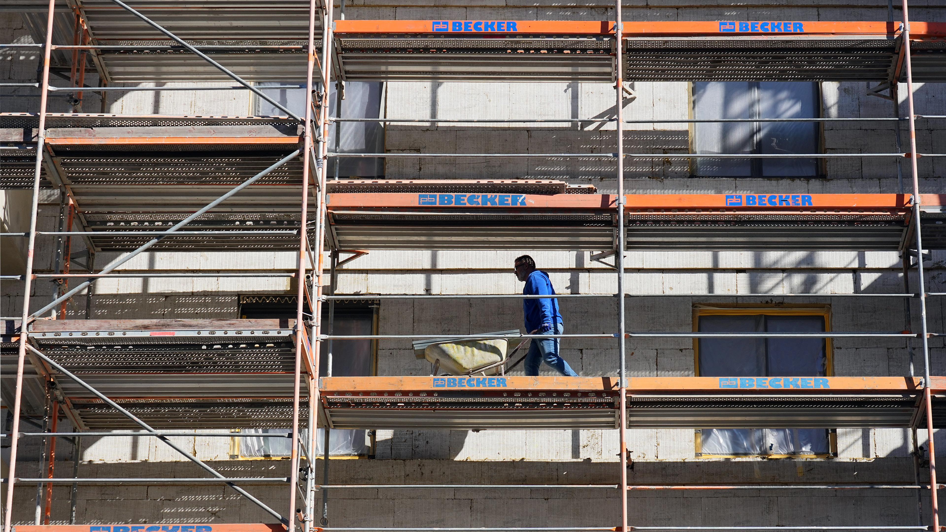 Ein Arbeiter geht auf einer Baustelle mit einer Schubkarre über das Baugerüst.