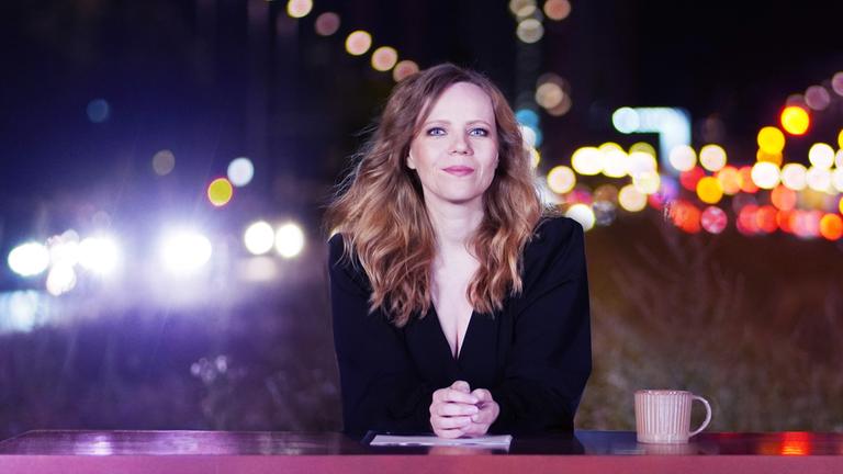 "Bosetti Late Night": Sarah Bosetti sitzt hinter einem pinkfarbenen Tresen. Sie schaut freundlich in die Kamera. Neben Ihr steht eine rosafarbene Kaffeetasse.