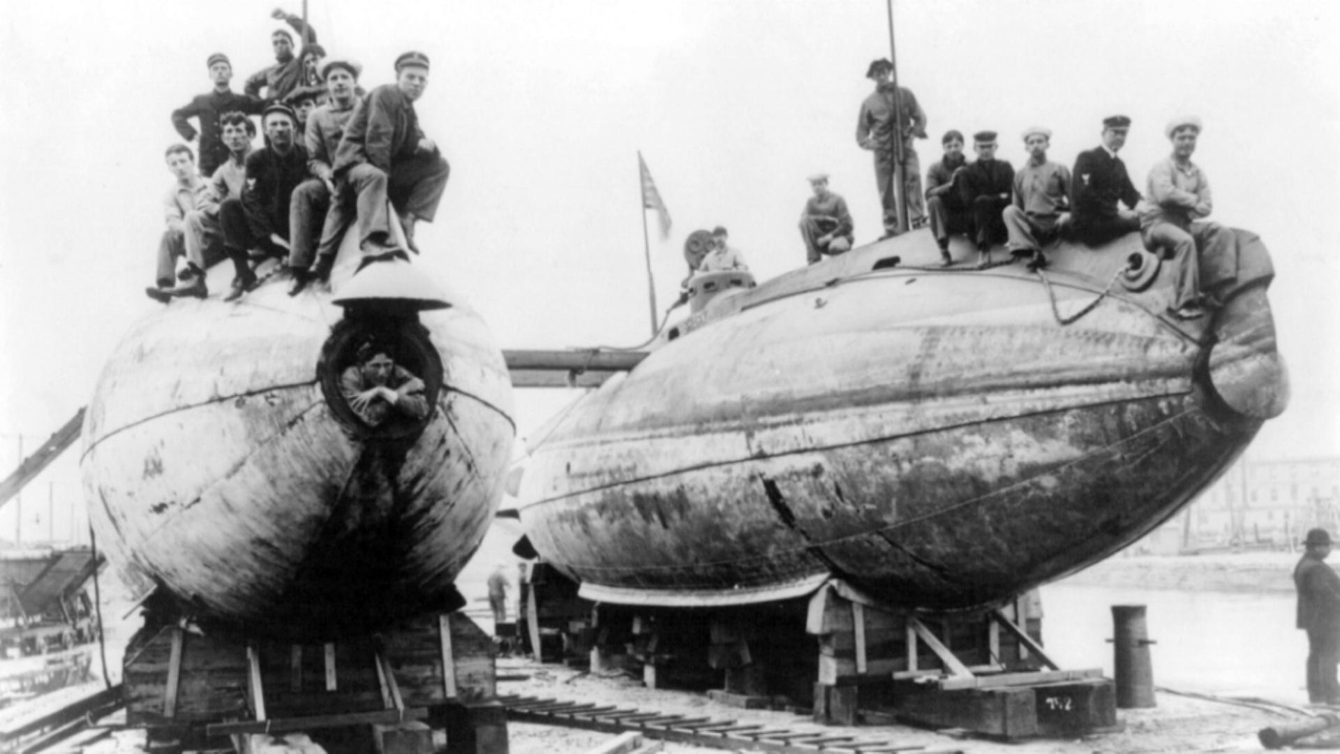 "U-Boote: Aufbruch in die Tiefe": Schwarz-Weiß-Aufnahme mit zwei großen U-Boot-Entwürfen, auf denen mehrere Menschen sitzen und für das Foto posieren.