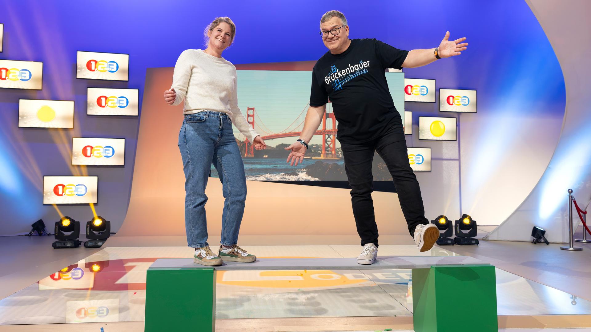 "1, 2 oder 3 - Über Brücken": Ingenieurin Anna Ostertag und Elton stehen im Studio auf einem Brett, dass auf zwei grünen Kästen liegt. Im Hintergrund befindet sich ein Bild einer Hängebrücke zwischen mehreren kleinen "1, 2 oder 3" - Logos.