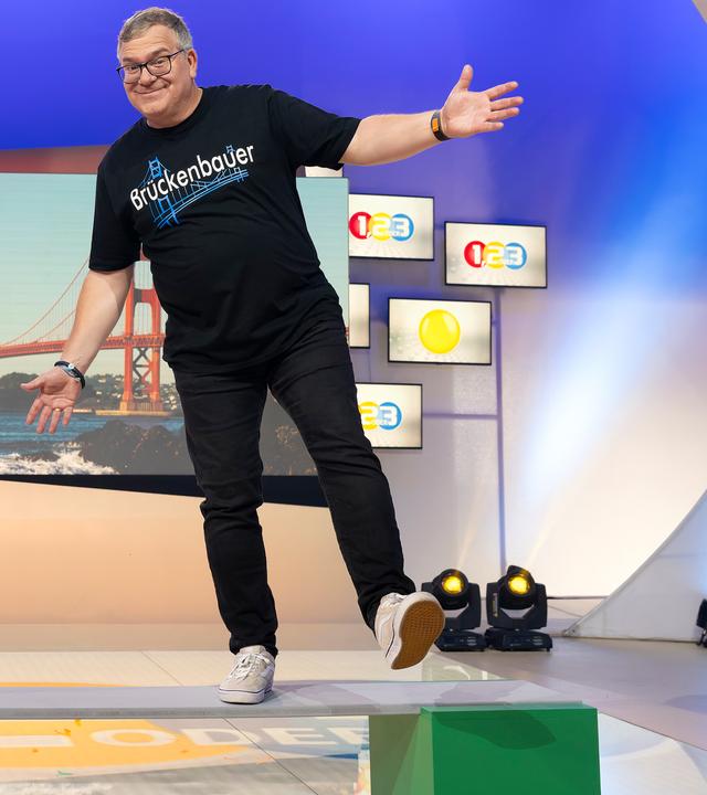 "1, 2 oder 3 - Über Brücken": Ingenieurin Anna Ostertag und Elton stehen im Studio auf einem Brett, dass auf zwei grünen Kästen liegt. Im Hintergrund befindet sich ein Bild einer Hängebrücke zwischen mehreren kleinen "1, 2 oder 3" - Logos.